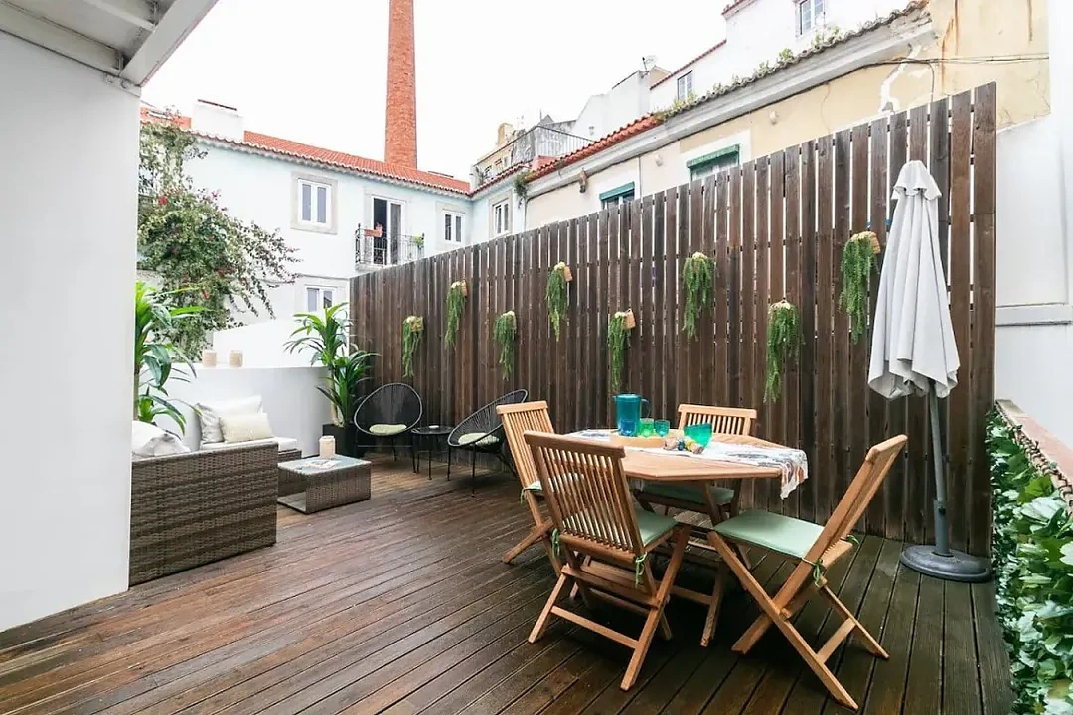 Um terraço com cerca de madeira e fetos pendurados, cadeirões de vime e uma mesa de refeições, com chaminés e telhados ao fundo