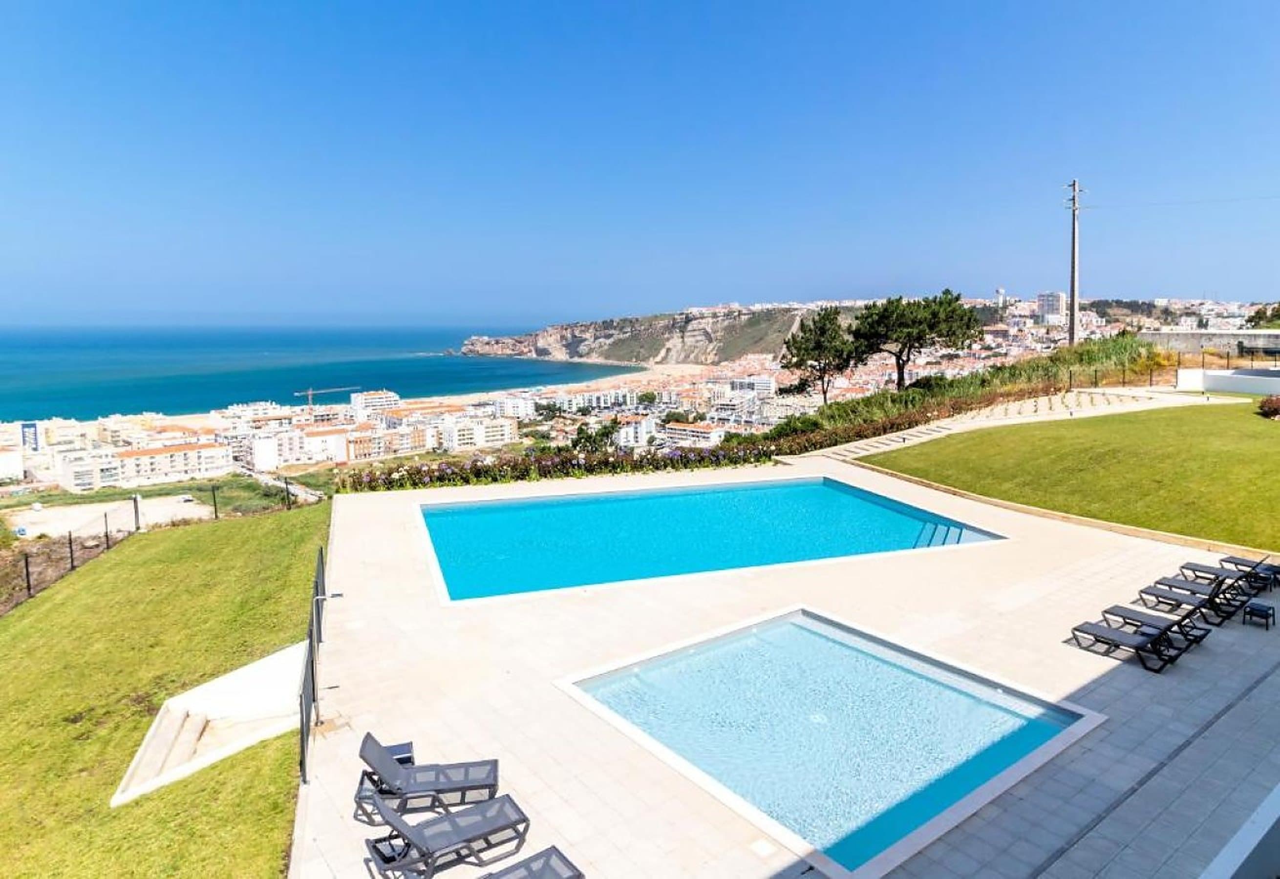 A hillside pool terrace looking down over the town of Nazaré, its bay and the cliff headland, with the Atlantic beyond