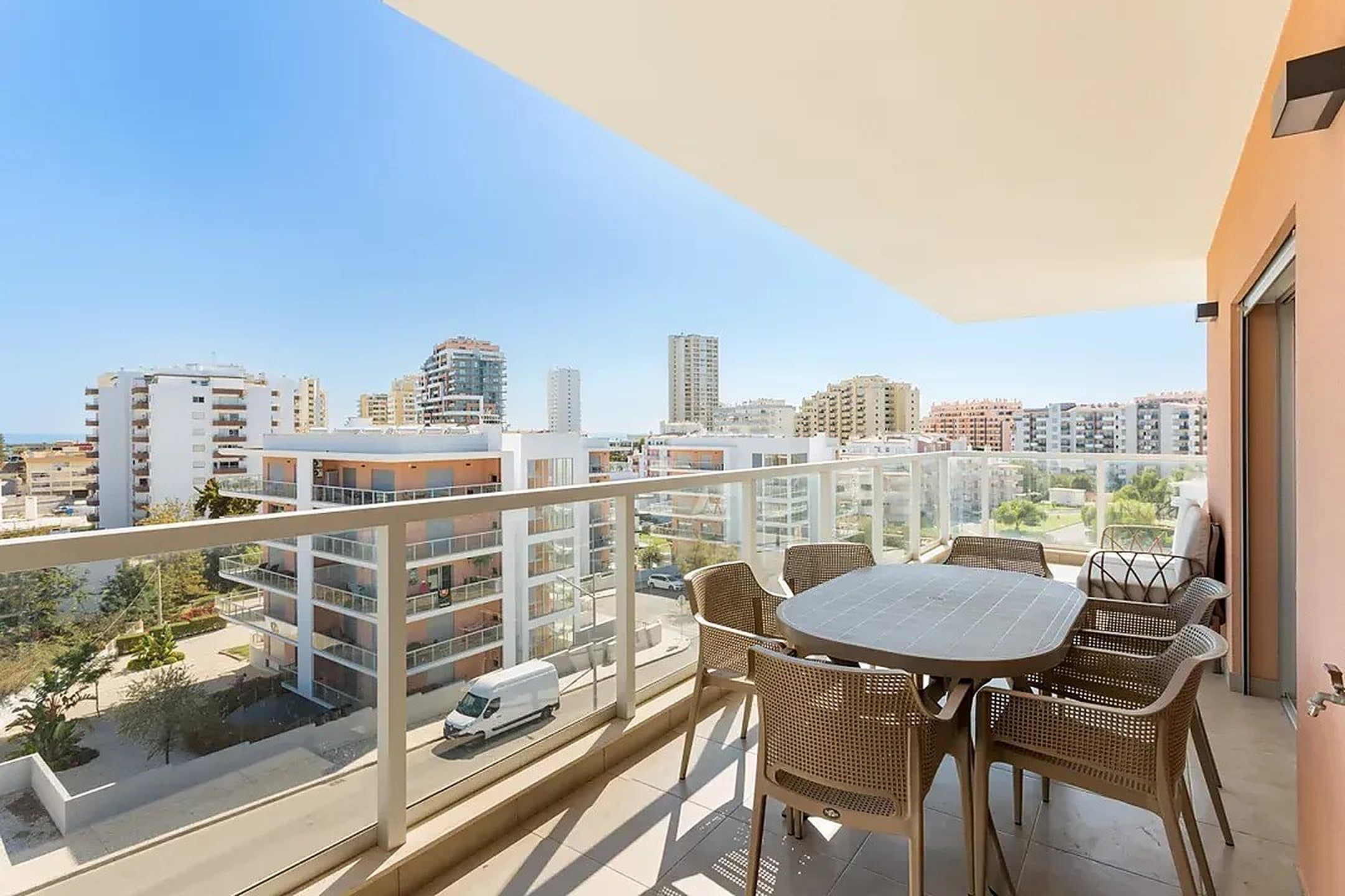 Uma varanda de um prédio alto com mobiliário de rattan sob uma vela de sombra, com vista para um conjunto de torres de apartamentos